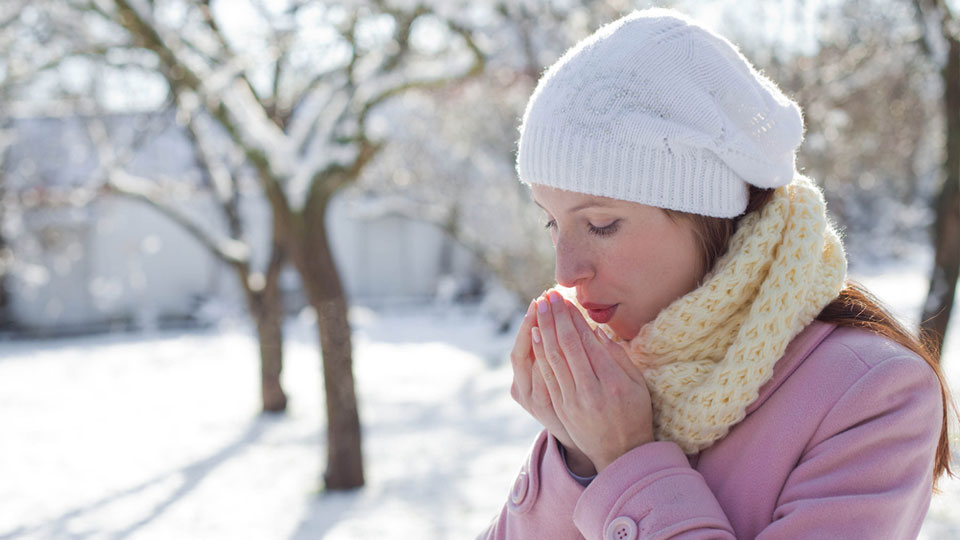 Cold Woman In Jacket, Cap and Scarf Warming Her Hands With Her Breath