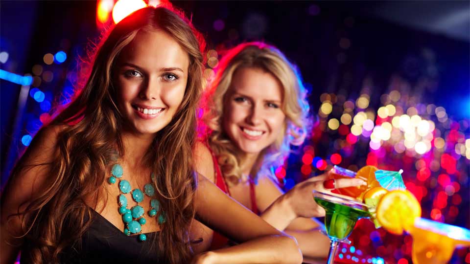 Two girls at a bar giving the camera flirtatious looks and enjoying brightly colored adult beverages